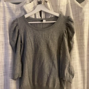 Grå stickad tröja med puffärm - Snygg grå stickad tröja med puffade trekvartsärmar och rund halsringning. Tröjan har ribbade muddar vid ärmslut och nederkant, vilket ger en schysst passform. Perfekt för dig som gillar enkel stil med en twist.