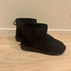 Svarta glittriga boots (uggs inspirerande) - Säljer ett par svarta boots med glittrig yta och platt sula. Skorna har rund tå och är fodrade med mjukt material på insidan för extra komfort. Perfekta för dig som vill ha lite extra bling i vardagen. Pris kan alltid diskuteras!💗