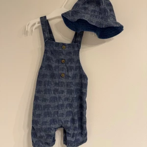 Blå hängselshorts med elefanter, 12-18 mån - Supersöta blå hängselshorts från Mothercare med elefantmönster och träknappar framtill. Gjorda i 100% bomull och har korsade axelband bak. Perfekt för varma dagar och kommer med matchande solhatt.