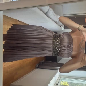 Champagnefärgad axelbandslös klänning - Säljer en champagnefärgad kortklänning med axelbandslös design och plisserad kjol. Midjan har ett brett band dekorerat med glittriga pärlor som ger extra lyxig känsla. Materialet är mjukt och har ett elegant fall, perfekt för dig som vill sticka ut.