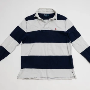 Randig långärmad skjorta Polo Ralph Lauren - Snygg långärmad skjorta från Polo Ralph Lauren med breda marinblå och vita ränder. Klassisk krage, knappar vid halsen och ikonisk röd logobrodyr på bröstet. Skjortan är gjord i mjuk bomull och har en relaxed fit som passar perfekt till jeans.