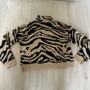 Beige och svart zebramönstrad polotröja - Snygg stickad polotröja med beige och svart zebramönster. Tröjan har lång ärm, ribbade muddar och hög krage.