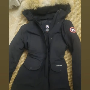 Svart dunjacka från Canada Goose (Trillium) - Säljer en svart dunjacka från Canada Goose med stor huva och äkta pälsdetalj. Jackan har två stora fickor framtill, klassisk logga på ärmen och dragkedja med knappar. Perfekt för kalla vinterdagar och riktigt snygg passform.