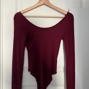 Vinröd långärmad bodysuit - Vinröd långärmad bodysuit med bred, rundad urringning och figurnära passform. Perfekt att styla med jeans eller kjol för en snygg look. Mjuk och stretchig känsla.