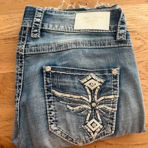 Blue Monkey Luxury jeans med broderad ficka - Snygga blå jeans från Blue Monkey Luxury med coola kontrastsömmar och en broderad bakficka med nitar och mönster. Jeansen har slitna detaljer och unika knappar med blommotiv. Perfekt för dig som gillar statement-plagg och detaljer.