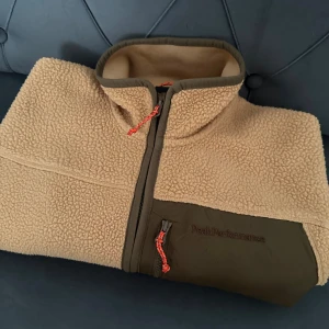Beige teddyjacka Peak Performance S - Säljer en beige teddyjacka från Peak Performance i storlek S. Jackan har mjuk fleece, detaljer i mörkgrönt tyg och orange snören på dragkedjorna. Hög krage och flera fickor med dragkedja gör den både snygg och praktisk. Perfekt för kyliga dagar. använd en gång
