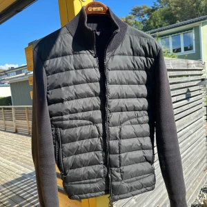 Svart quiltad pufferjacka Moncler - Svart pufferjacka från Moncler med quiltad framsida och stickade ärmar. Jackan har ribbad krage, dragkedja framtill och Moncler-logga på ärmen. Snygg, stilren design som passar perfekt till höst och vinter. Materialmix av polyester och stickad bomull. Pris kan diskuteras 