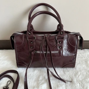 Brun handväska med dragkedja - Snygg handväska i skinn med coola detaljer som silverfärgad dragkedja och långa remmar framtill. Väskan har både handtag och avtagbar axelrem, perfekt för att bära på olika sätt. Stilren design med metallnitar och struktur i materialet.