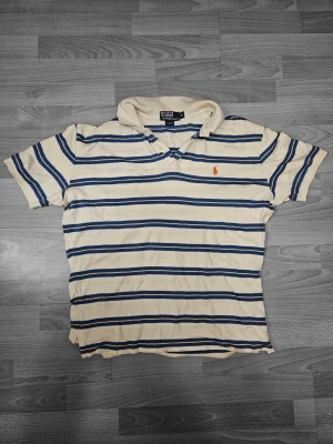 Randig pikétröja från Ralph Lauren M - Klassisk pikétröja från Polo Ralph Lauren i beige med blå och gula ränder. Tröjan har korta ärmar, krage och knappslå framtill. Broderad röd logga på bröstet. 