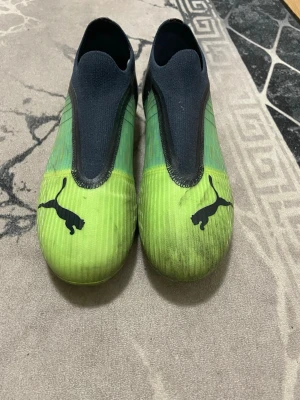 Puma Ultra fotbollsskor grön/svart - Säljer ett par Puma Ultra fotbollsskor i grön och svart med coolt grafiskt mönster och tydlig Puma-logga på tån. Skorna har en strumpliknande ovandel utan snörning och platt sula med dobbar för gräsplan. Perfekt för dig som vill ha speed och stil på planen.