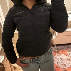 Mörkblå dunjacka från Peak Performance - Säljer en svart dunjacka från Peak Performance med quiltad design och dragkedja framtill. Jackan har en figurnära passform och huva, perfekt för kyliga dagar. Lätt och smidig att bära, passar till det mesta och är enkel att styla.