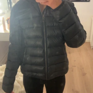 Colmar pufferjacka - Säljer en svart/mörkblå pufferjacka från Colmar Originals med klassisk logga på ärmen. Jackan har en rak passform, långa ärmar och grova horisontella sömmar. Dragkedja framtill och lätt glansigt material som ger en snygg look. Perfekt för kyliga dagar. (Finns defekt på ena ärmen, sista bilden. Men syns knappt när den används och går lätt att sy!)