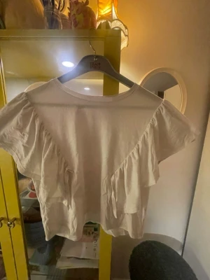 Vit blus med volanger från ärmen till naveln. Stl L - Den är i bra skick, lite kort till magen. Den har ett mjukt meterial och passar till det mesta.