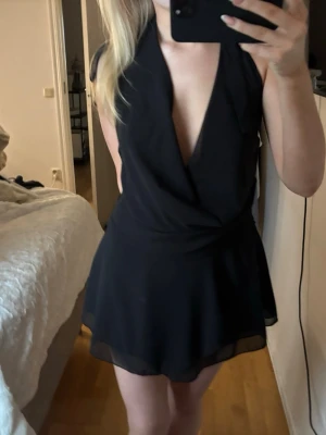 Svart omlottklänning med öppen rygg - Säljer en svart kortklänning med omlottdesign och djup v-ringning. Klänningen har tunna band som knyts i nacken och en snygg öppen rygg. Kjolen är lätt utsvängd och tyget känns luftigt och faller fint.