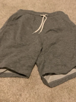 Grå mjukisshorts från Next med snörning - Grå mjukisshorts från Next med vit snörning i midjan och regular fit. Tillverkade i mjuk bomullsblandning (90% bomull, 10% viskos) som känns skön mot huden. Perfekta för chill dagar eller träning.