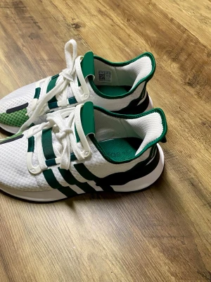 Vita och gröna adidas sneakers, saiz 40/5 - Säljer ett par fräscha adidas sneakers i vitt och grönt med klassiska tre ränder på sidan. Skorna har snörning, mesh-överdel och en grå sula med bra grepp. Insidan är grön och detaljerna är riktigt cleana. Perfekta för dig som gillar sportig stil.