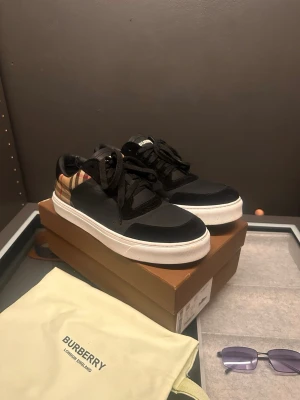 Burberry sneakers  - Snygga Burberry sneakers i svart med klassiskt beige och röd-rutigt mönster på hälen. Skorna har vita, platta sulor och svarta snören. Tillverkade i en mix av skinn och textil för en lyxig känsla. Perfekta för dig som vill ha en stilren men ändå statement look.