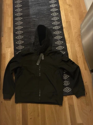 Svart vindjacka från Stone Island - Säljer en svart vindjacka från Stone Island med huva och dragkedja framtill. Jackan har den klassiska loggan på vänster ärm och praktiska fickor. Materialet är polyester och passformen är normal. Perfekt för dig som vill ha en clean och stilren look.