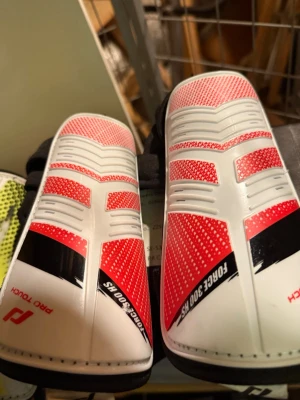 Vita och röda benskydd Pro Touch - Säljer ett par benskydd från Pro Touch i vitt och rött med svarta detaljer. De har en sportig design med mönstrade ytor och texten FORCE 300 HS. Perfekta för dig som spelar fotboll och vill ha bra skydd. 