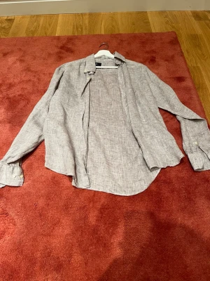 Ljusgrå linneskjorta Hanen & Jacob  - Säljer en stilren ljusgrå skjorta från Hansen & Jacob i luftigt linne. Skjortan har klassisk krage, långa ärmar och knappar framtill samt vid ärmsluten. Perfekt för en chill och avslappnad look.