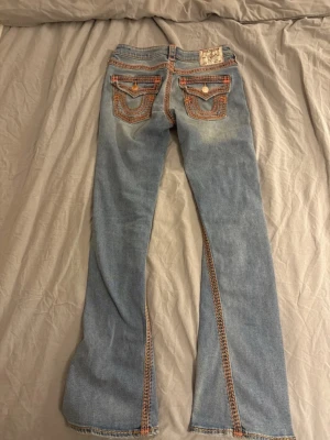 True Religion ljusblå bootcut jeans - Säljer ett par ljusblå jeans med orangerosa broderi runt om byxor från True religon. Modellen på jeansen joey low Rise och flare, i storlek 26. Köpte jeansen på Nelly för nypris 2300 men de passade inte så jag säljer de här nu.