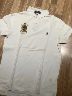 Vit pikétröja Polo Ralph Lauren S - Vit pikétröja från Polo Ralph Lauren i bomull, custom fit. Klassisk krage, kortärmad och med broderad logga samt emblem på bröstet. Perfekt för en clean och sportig stil.