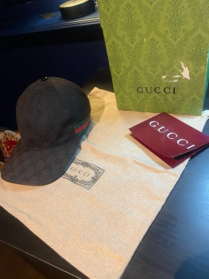 Svart Gucci keps - Säljer denna feta svarta gucci kepsen med klassiskt GG-mönster och grönt/rött band på sidan. Kepsen har böjd skärm och justerbar passform. Perfekt för dig som vill ha en stilren accessoar med lyxig känsla. Lite solblekt som ni kan se på bilderna därav prissänkning annars som ny. Original box, kvitto och dustbag ingår