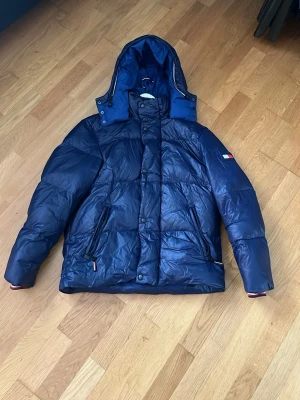 Blå pufferjacka från Tommy Hilfiger - Säljer en blå pufferjacka från Tommy Hilfiger i storlek M. Jackan har huva, dragkedja och knappar framtill samt två fickor med dragkedja. Klassisk Hilfiger-flagga på ärmen och insidan. Perfekt för kalla dagar och riktigt snygg streetstil.