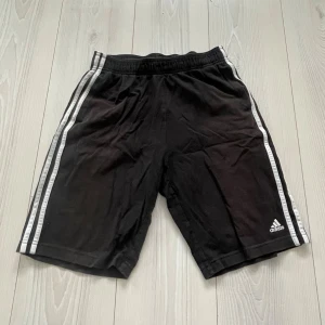 Svarta Adidas shorts  - Säljer ett par klassiska svarta Adidas shorts med vita ränder längs sidorna och Adidas-logga på vänster ben. De har elastisk midja och är gjorda i ett lätt och sportigt material, perfekta för träning eller chill. Använda väldigt lite så otroligt mycket bra skick, storlek S! Vid intresse bara att höra av sig. 
