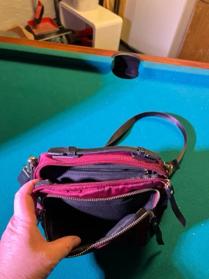 Mörkröd axelväska med flera fack - Säljer en mörkröd axelväska med justerbar rem och flera praktiska dragkedjefack. Väskan har detaljer i svart och guld, och är tillverkad i ett glansigt syntetmaterial. Perfekt storlek för att få plats med allt du behöver och enkel att bära över axeln.