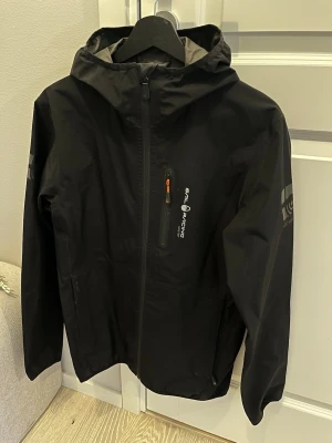 Svart Gore-Tex jacka från Sail Racing - Svart vindjacka från Sail Racing med Gore-Tex-material och huva. Jackan har dragkedja framtill, en bröstficka med orange detalj och logga på bröstet. Perfekt för blåsiga och regniga dagar, med stilren design och funktionella detaljer.