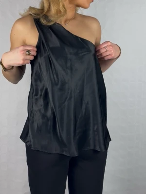 Svart oneshoulder satin-topp - Snygg svart topp i satin med oneshoulder-design. Toppens glansiga material ger en lyxig känsla och den har en lös passform som faller snyggt. Perfekt för dig som gillar stilrena och trendiga plagg med en twist.
