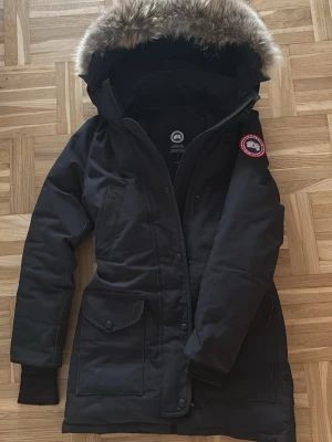 Svart dunjacka från Canada Goose - Säljer en svart Canada Goose dunjacka med stor huva och fluffig päls som går att ta av. Jackan har flera fickor, dragkedja och knappar framtill samt klassisk logga på ärmen. Perfekt för kalla vinterdagar och riktigt snygg passform.