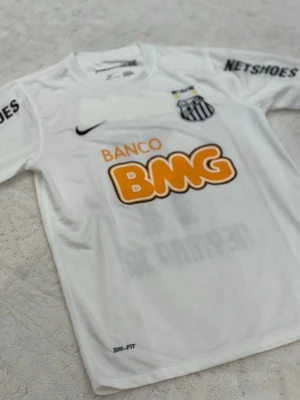 Vit Santos FC Neymar Jr tröja - Säljer en vit Santos FC matchtröja från Nike med Neymar Jr och nummer 11 på ryggen. Tröjan har svarta och orange tryck, Dri-Fit material och flera sponsorloggor. Perfekt för dig som älskar fotboll och vill ha en ikonisk supportertröja.