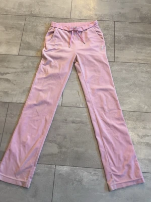 Rosa velourbyxor från Juicy Couture - Säljer ett par ikoniska ljusrosa mjukisbyxor från Juicy Couture i mjuk velour. Byxorna har normal passform, raka ben och snörning i midjan. På baksidan står det 'Juicy' i glittriga stenar. Perfekta för en chill och trendig look.