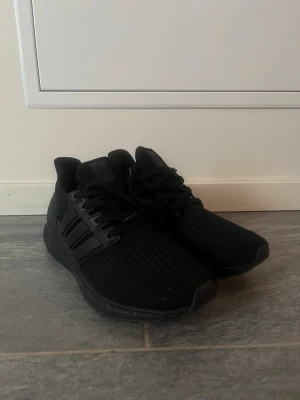 Svarta Adidas Ultraboost sneakers - Säljer ett par helt nya helsvarta Adidas Ultraboost sneakers med stickad ovandel och klassiska tre ränder på sidan. Skorna har snörning, platt sula och en riktigt skön boost-dämpning. Perfekta för dig som vill ha både stil och komfort i vardagen.