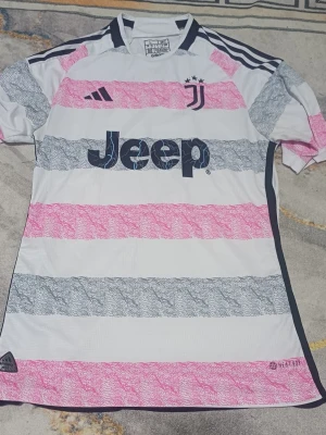 Juventus Adidas matchtröja 2023 L - Juventus officiella matchtröja från Adidas, säsong 2023. Tröjan är vit med breda rosa och grå ränder, svart Juventus-logga och Jeep-tryck på bröstet. Slim fit-modell tillverkad av återvunnen polyester (Parley Ocean Plastic). Autentisk badge nertill.