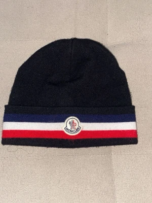 Moncler mössa med ränder - Snygg svart Moncler mössa med breda ränder i blått, vitt och rött runt kanten. Klassisk Moncler-logga framtill. Stickad i mjukt material som håller dig varm och passar perfekt till vintern.