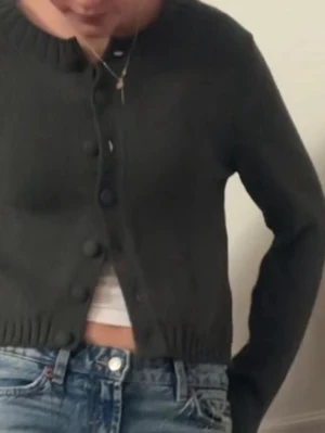 Mörkgrå kofta med knappar - Snygg mörkgrå kofta med rund hals och stora knappar framtill. Modellen är croppad och har långa ärmar samt ribbade muddar vid ärmslut och nederkant. Perfekt att slänga över en t-shirt till jeans!🔥