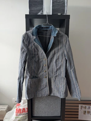 Randig jeanskavaj från Prêt à Porter - Snygg randig kavaj i jeansmaterial från Prêt à Porter, storlek 44. Jackan har blå denimkrage, dekorativa knappar framtill och två stora fickor. Baksidan har en denimdetalj med knappar för extra stilpoäng.