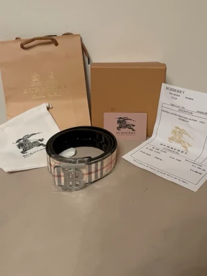 Vändbart bälte (beige/svart) med silverlogga - Stilrent och exklusivt bälte med vändbar design. Ena sidan är beige med klassiskt mönster och den andra sidan svart, vilket gör det enkelt att matcha till olika outfits. Silverfärgad logga ger en clean och lyxig känsla. Passar perfekt till både vardag och finare tillfällen.