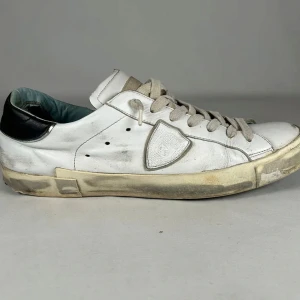 Vita sneakers i skinn med svart detalj - Säljer ett par vita sneakers i skinn med svart detalj på hälen och grå snörning. Skorna har en klassisk låg profil, rund tå och platt sula. Sidan pryds av en sköldformad patch. Perfekta för dig som gillar en avslappnad och trendig look.