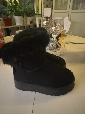 Svarta boots med fuskpäls och rosetter - Säljer ett par svarta boots med fluffig fuskpäls runt skaftet och söta rosetter bak. Skorna har en platt, rejäl sula och är gjorda i mockaimitation. Perfekta för kalla dagar när du vill vara både varm och stylish. Storleken känns som en 35 inte 36 men det står 36 