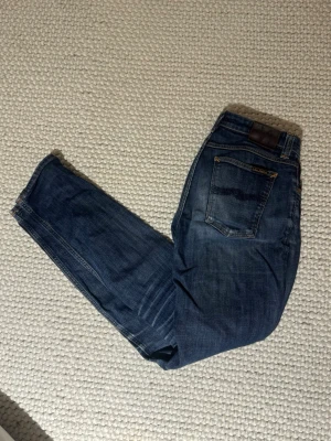 MörkBlå jeans från Nudie  - Säljer ett par klassiska mörkblå jeans från Nudie med snygga slitningar och kontrastsömmar. Jeansen har rak passform och fem fickor, perfekta till sneakers eller boots. Materialet är slitstarkt denim i bomull. Notera att Gylfen har en liten defekt o måste fixas, därav pris