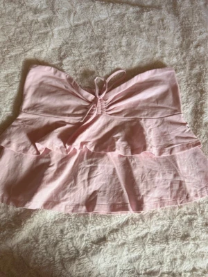 Ljusrosa volangtop från Gina Tricot - Supersöt ljusrosa topp från Gina Tricot med volangdetaljer och knyt framtill. Toppen är ärmlös och har en luftig passform, perfekt för varma dagar. Tillverkad i mjuk bomull som känns skön mot huden. Strl XL! 