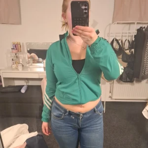 Grön Adidas jacka - Grön typ vindjacka från Adidas med svarta detaljer på axlarna och vita ränder på ärmarna. Tröjan har dragkedja framtill, lång ärm och klassisk Adidas-logga på bröstet.