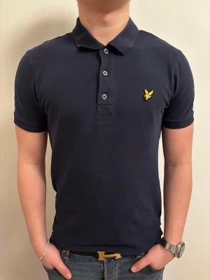 Mörkblå pikétröja från Lyle & Scott - Säljer en stilren mörkblå pikétröja från Lyle & Scott med klassisk krage och tre knappar framtill. Tröjan har korta ärmar och ett gult broderat logomärke på bröstet. Perfekt till jeans eller shorts för en clean look. Kom privat för att diskutera pris. 100% äkta