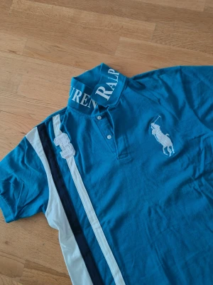Blå pikétröja från Ralph Lauren - Säljer en blå pikétröja från Ralph Lauren med vit och marinblå rand på vänster sida, stor vit logga på bröstet och broderad siffra 3. Kragen har stor text med märkesnamnet. Klassisk piké med korta ärmar och knappar vid halsen.