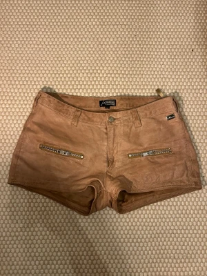Bruna skinnshorts - Snygga bruna shorts från Kenza i fakeskinn med två dragkedjor framtill och bakfickor. De har bälteshällor och en lågmidjad passform. 