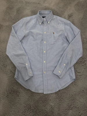 Ljusblå skjorta från Ralph Lauren XS - Klassisk ljusblå skjorta från Ralph Lauren i bomull. Skjortan har button-down krage, långa ärmar och vita knappar framtill. Ikoniska broderade loggan på bröstet ger en clean och tidlös look. Perfekt för dig som gillar stilrena plagg med snygg passform.